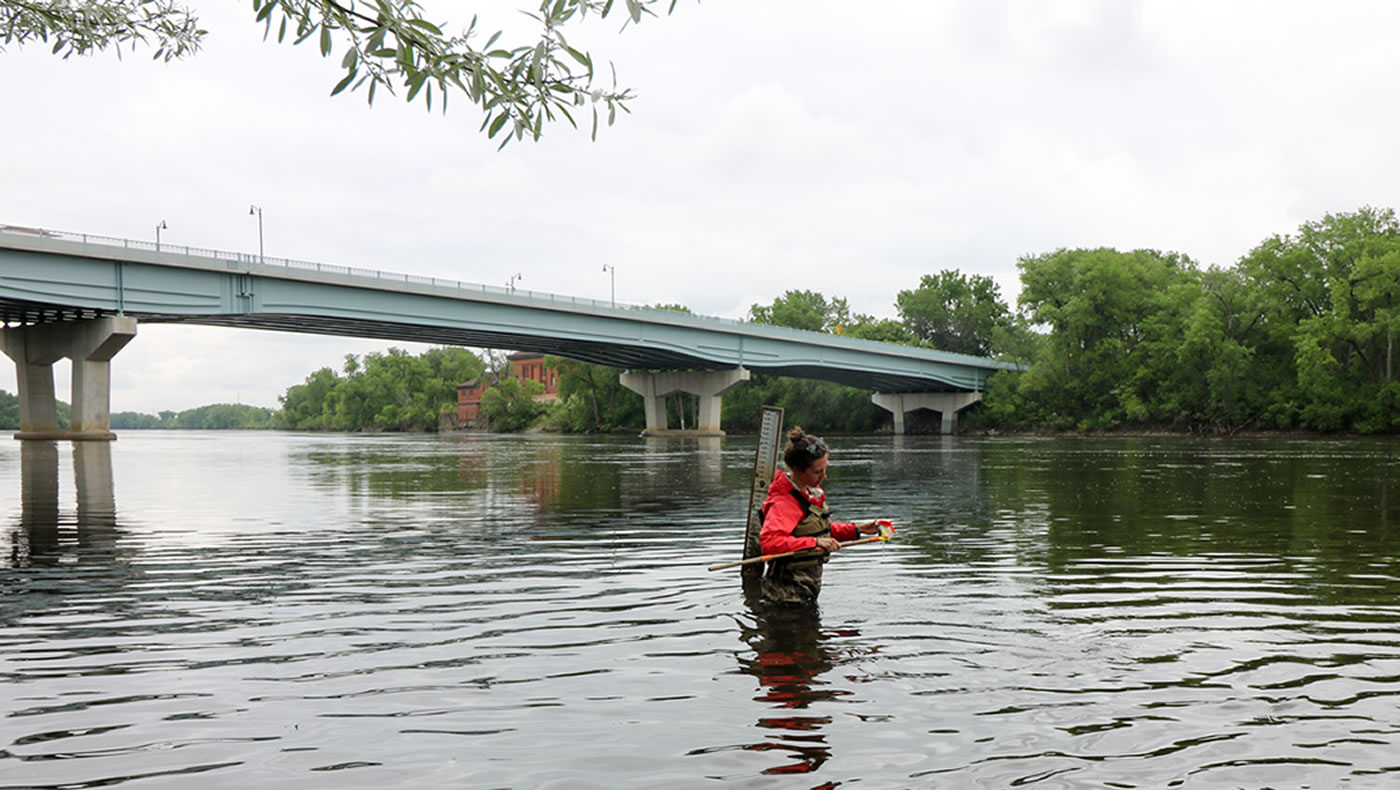 MWMO monitoring team member wades into the Mississippi River to check water levels and gather a sample by way of a bottle attached to the end of a wooden pole to avoid any contact with hands, gear, or anything else that could contaminate the sample. 