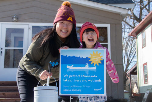 A woman and child pose with an Adopt-A-Drain lawn sign.