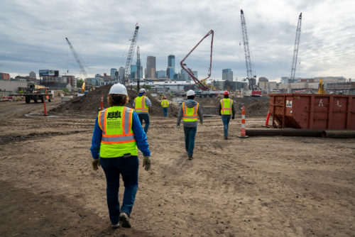 Workers walk toward a construction site with downtown Minneapolis in the background.