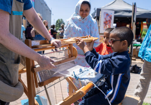 Children practice using a loom at an outdoor Weaving Water fiber art event.