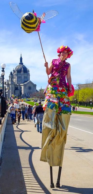 Tara Fahey on stilts walks across a bridge carrying a large bee lanter on a pole. They wear a colorful floral costume, and a Cathedral rises in the background beneath a blue sky.