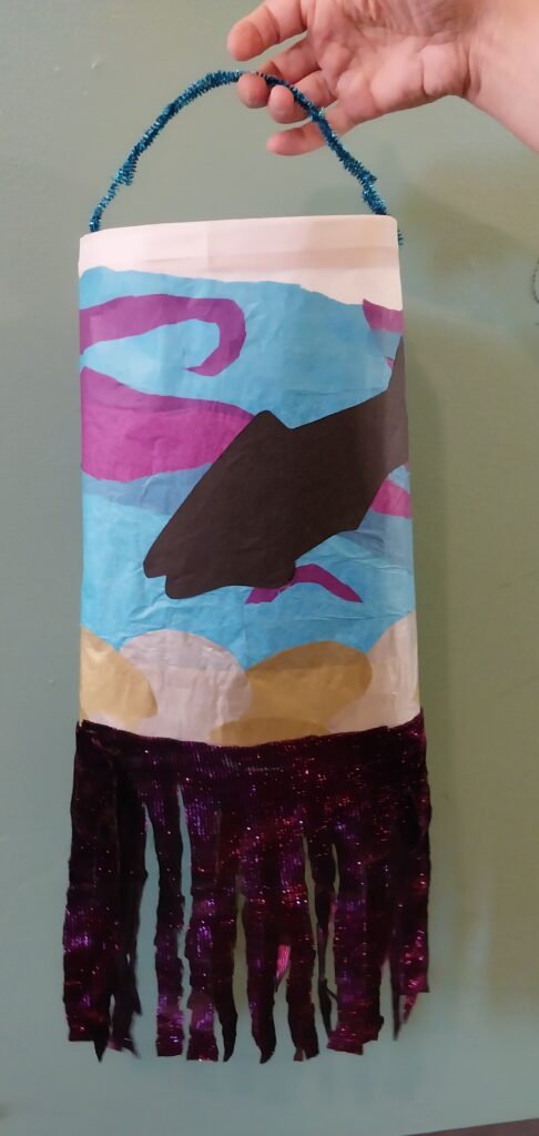 Handmade paper lantern decorated with a fish silhouette swimming above a riverbed. Blue and purple paper creates a water scene, with shimmering purple streamers hanging from the bottom and a blue handle at the top