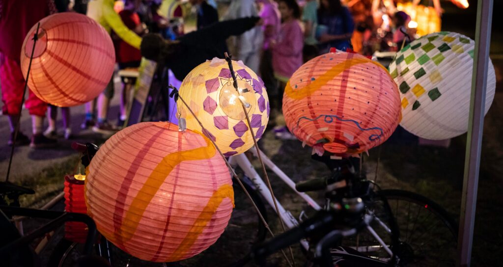Several illuminated paper lanterns decorated with colorful patterns are attached to bicycles during an evening community event. People gather in the softly lit background.
